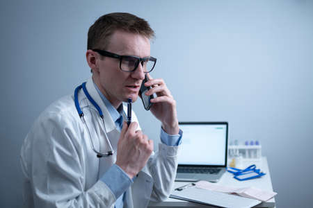 European doctor in white medical coat and glasses consults patient on cell phone in clinic office, sitting at table with laptop. German general practitioner at workplace in hospital talking on phoneの写真素材
