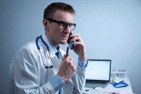 Smiling doctor having phone call at desk. Physicianr talking with his patient in office clinic sitting at table with laptop. German doctor in white lab coat works hospital, consults online by phoneの写真素材