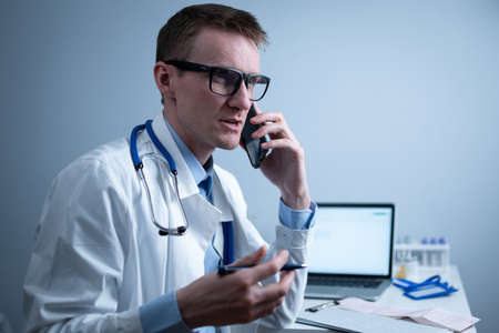 European doctor in white medical coat and glasses consults patient on cell phone in clinic office, sitting at table with laptop. German general practitioner at workplace in hospital talking on phoneの写真素材