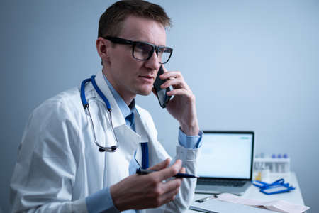 European doctor in white medical coat and glasses consults patient on cell phone in clinic office, sitting at table with laptop. German general practitioner at workplace in hospital talking on phoneの写真素材