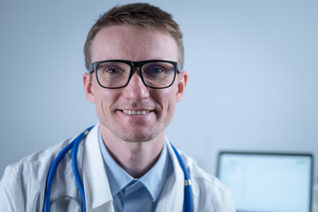 Portrait of adult handsome family doctor in medical office. Successful physician in white lab coat looking at camera and smiling in hospital clinic. Young general practitioner wearing glasses smilingの写真素材