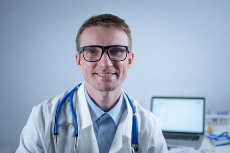 Portrait of adult handsome family doctor in medical office. Successful physician in white lab coat looking at camera and smiling in hospital clinic. Young general practitioner wear glasses smiling.の写真素材