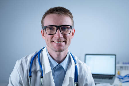 Portrait of adult handsome family doctor in medical office. Successful physician in white lab coat looking at camera and smiling in hospital clinic. Young general practitioner wearing glasses smilingの写真素材
