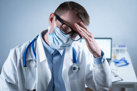 Exhausted young doctor in lab coat takes off mask and glasses, rubs eyes, has headache from fatigue and overexertion from working in heavy shift in hospital. Doctor looks tiredly at camera in officeの写真素材
