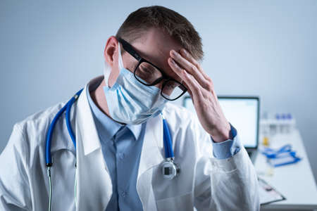 German European young doctor looking at camera wearing glasses and mask in clinic office at workplace with tired look. Topic is the hard work of medical workers during the coroavirus pandemicの写真素材