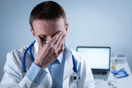 German European young doctor looking at camera wearing glasses and mask in clinic office at workplace with tired look. Topic is the hard work of medical workers during the coroavirus pandemicの写真素材