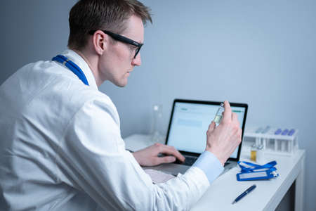 Scientist in medical laboratory examines vial of medicine, vaccine against viruses, influenza, covid. Doctor enters information into computer about investigation. Immunization from terrible diseasesの写真素材