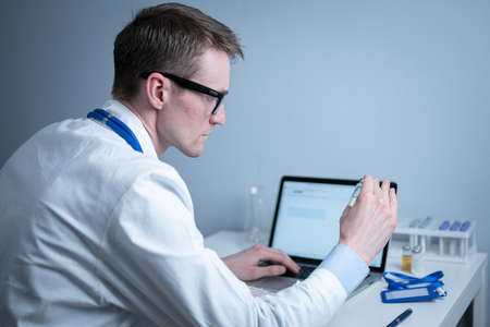 Concept of medicine and immunization of human body against infections, viruses to prevent epidemics of diseases. Scientist examines vial of medicine and works on laptop in medical laboratory. Vaccineの写真素材