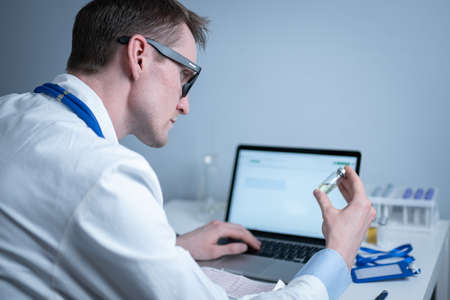 Scientist in medical laboratory examines vial of medicine, vaccine against viruses, influenza, covid. Doctor enters information into computer about investigation. Immunization from terrible diseasesの写真素材