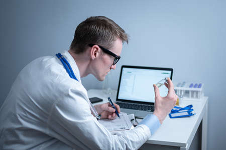 Young caucasian doctor working at table in hospital laboratory examining an ampoule of medicine and entering data into computer. New vaccine against coronavirus. Scientist changing future covid 19の写真素材