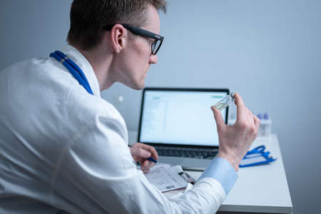 Scientist in medical laboratory examines vial of medicine, vaccine against viruses, influenza, covid. Doctor enters information into computer about investigation. Immunization from terrible diseasesの写真素材