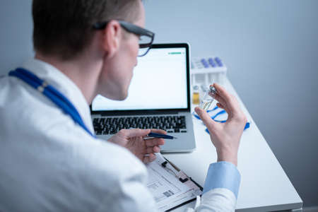 Young caucasian doctor working at table in hospital laboratory examining an ampoule of medicine and entering data into computer. New vaccine against coronavirus. Scientist changing future covid 19の写真素材