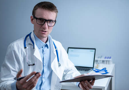 Young male doctor consults patient in hospital office, holding medical examinations in hands. General practitioner in white medical coat looks into camera and makes an appointment with patient onlineの写真素材