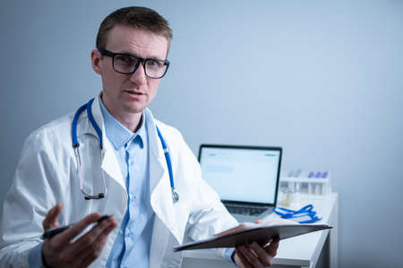 Young male doctor consults patient in hospital office, holding medical examinations in hands. General practitioner in white medical coat looks into camera and makes an appointment with patient onlineの写真素材