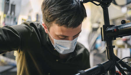 Bicycle shop repairman works in bicycle service and repair workshop during coronavirus quarantine wearing face shield and gloves, new norm. Mechanic repairing bicycle wheel wearing medical maskの写真素材