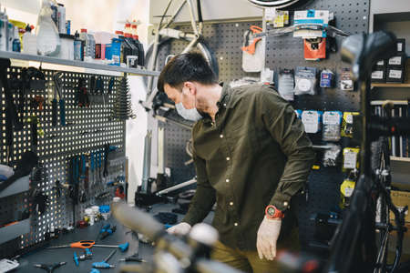 Bicycle mechanic wearing protective mask and gloves repairs customers bicycle wheel in accordance with quarantine standards during coronavirus pandemic. Small business during covid 19 lockdownの写真素材