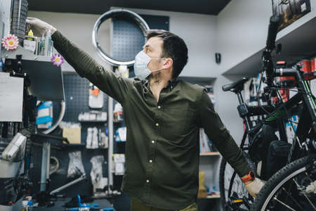 Mechanic repairs bicycle in workshop wearing covid 19 mask. Worker of bicycle store fixes bike during coronavirus quarantine in protective mask. Repairman maintenance cycle. Sport shop conceptの写真素材