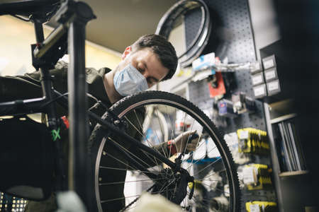 Bicycle mechanic wearing protective mask and gloves repairs customers bicycle wheel in accordance with quarantine standards during coronavirus pandemic. Small business during covid 19 lockdownの写真素材