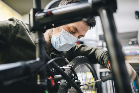 Bicycle shop repairman works in bicycle service and repair workshop during coronavirus quarantine wearing face shield and gloves, new norm. Mechanic repairing bicycle wheel wearing medical maskの写真素材