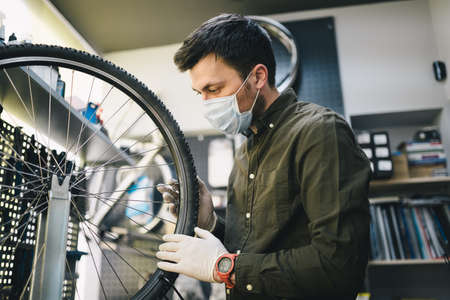 Bicycle mechanic wearing protective mask and gloves repairs customers bicycle wheel in accordance with quarantine standards during coronavirus pandemic. Small business during covid 19 lockdownの写真素材