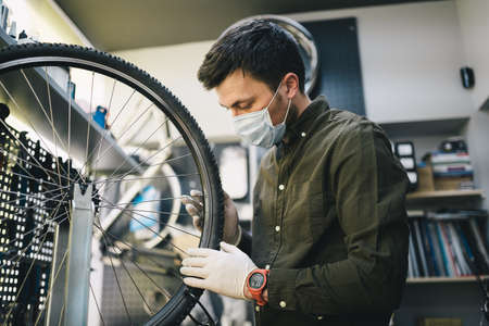 Bicycle shop repairman works in bicycle service and repair workshop during coronavirus quarantine wearing face shield and gloves, new norm. Mechanic repairing bicycle wheel wearing medical maskの写真素材