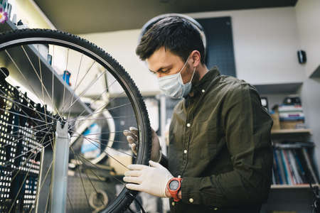 Mechanic repairs bicycle in workshop wearing covid 19 mask. Worker of bicycle store fixes bike during coronavirus quarantine in protective mask. Repairman maintenance cycle. Sport shop conceptの写真素材