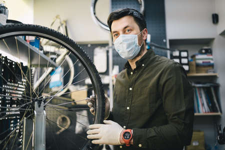 Bicycle mechanic wearing protective mask and gloves repairs customers bicycle wheel in accordance with quarantine standards during coronavirus pandemic. Small business during covid 19 lockdownの写真素材