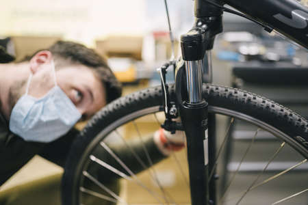 Bicycle mechanic wearing protective mask and gloves repairs customers bicycle wheel in accordance with quarantine standards during coronavirus pandemic. Small business during covid 19 lockdownの写真素材