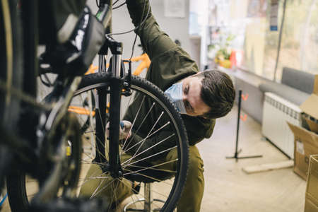 Bicycle mechanic wearing protective mask and gloves repairs customers bicycle wheel in accordance...