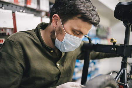 Bicycle shop repairman works in bicycle service and repair workshop during coronavirus quarantine wearing face shield and gloves, new norm. Mechanic repairing bicycle wheel wearing medical maskの写真素材