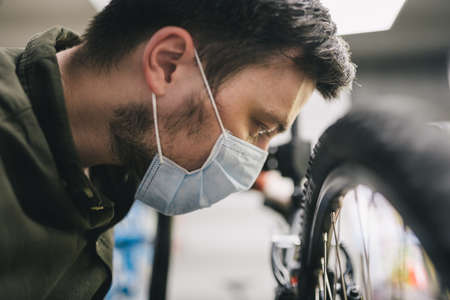 Bicycle mechanic wearing protective mask and gloves repairs customers bicycle wheel in accordance with quarantine standards during coronavirus pandemic. Small business during covid 19 lockdownの写真素材