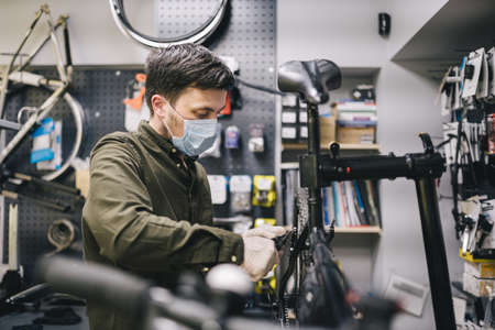 Mechanic repairs bicycle in workshop wearing covid 19 mask. Worker of bicycle store fixes bike during coronavirus quarantine in protective mask. Repairman maintenance cycle. Sport shop conceptの写真素材