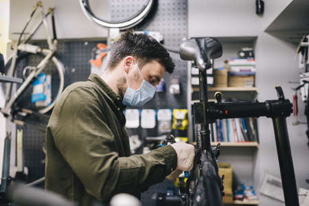 Bicycle mechanic wearing protective mask and gloves repairs customers bicycle wheel in accordance with quarantine standards during coronavirus pandemic. Small business during covid 19 lockdownの写真素材