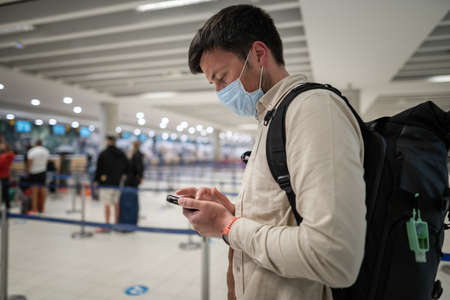 Male passenger in mask with backpack uses smartphone and stands with social distance on check-in line at airport of Cyprus, paphos city. Travel by plane during quarantineの写真素材