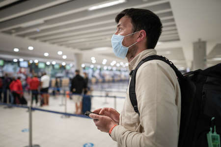 Man in protective mask with phone at airport departure check-in desk for baggage drop-off at terminal and check-in for flight, maintains social distance.の写真素材
