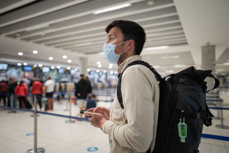 Flight rules during  pandemic only in protective face mask. New normal concept. Masked man with phone in hand and backpack stand in line to check in for plane. Social distancing in airoportの写真素材
