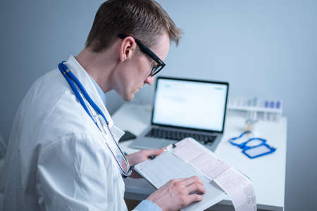 Young handsome doctor man in glasses and white lab coat examines results of electrocardiographic...