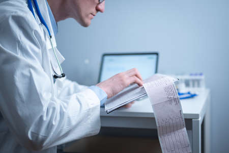 Cardiologist reading an ECG print-out. Doctor analyzing electrocardiogram. Practitioner examine patient test results. Medical and healthcare concept. Physician looking at cardiogram at medical officeの写真素材