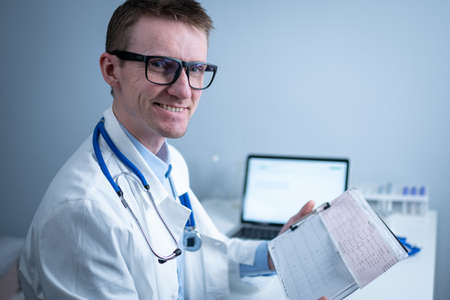 Young handsome doctor man in glasses and white lab coat examines results of electrocardiographic examination of patient heart in hospital office. Cardiologist looking at cardiogram at medical officeの写真素材