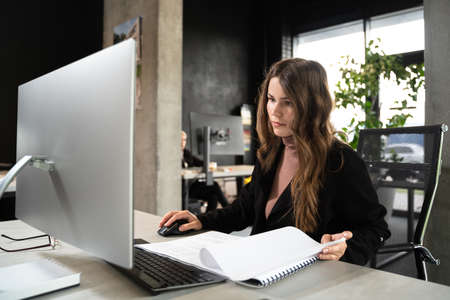 Young woman architect working with blueprints and technical design project at work table and computer in office. Designer using architectural plans of buildings, working at computer, creates projectの写真素材
