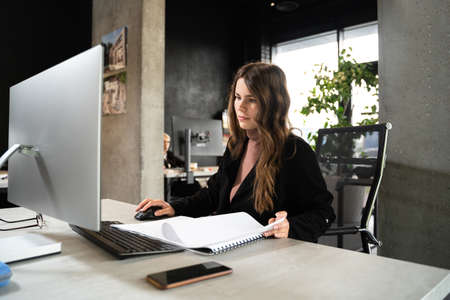 Young woman architect working with blueprints and technical design project at work table and computer in office. Designer using architectural plans of buildings, working at computer, creates projectの写真素材