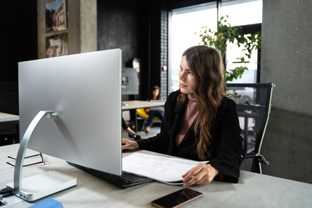 Young woman architect working with blueprints and technical design project at work table and computer in office. Designer using architectural plans of buildings, working at computer, creates projectの写真素材