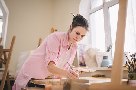 Caucasian woman artist working on a painting in bright daylight studio. Happy artist draws an art project with paints and a brush in the workshop. Hobby. Artist at work. Creative profession.の写真素材