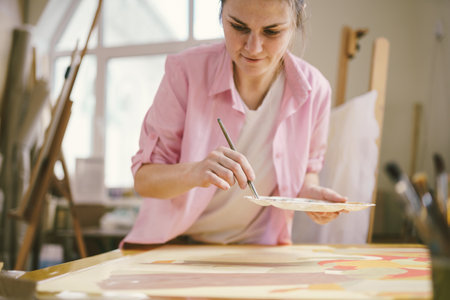 Caucasian woman artist working on a painting in bright daylight studio. Happy artist draws an art project with paints and a brush in the workshop. Hobby. Artist at work. Creative profession.の写真素材