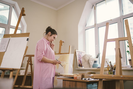 Cute woman paints on canvas in an art workshop. Artist creating picture. Art school or studio. Work with paints, brushes and easel. Hobby and leisure concept. Woman painter at workspace.の写真素材