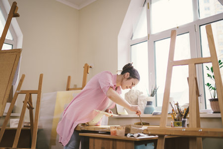 Caucasian woman artist working on a painting in bright daylight studio. Happy artist draws an art project with paints and a brush in the workshop. Hobby. Artist at work. Creative profession.の写真素材