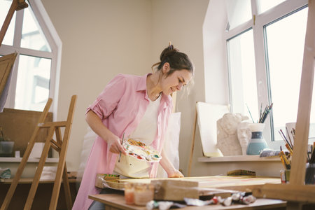Cute woman paints on canvas in an art workshop. Artist creating picture. Art school or studio. Work with paints, brushes and easel. Hobby and leisure concept. Woman painter at workspace.の写真素材