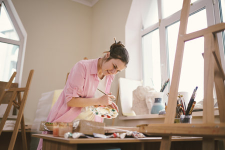 Caucasian woman artist working on a painting in bright daylight studio. Happy artist draws an art project with paints and a brush in the workshop. Hobby. Artist at work. Creative profession.の写真素材