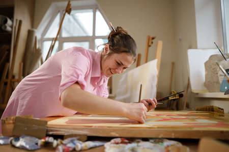 Caucasian woman artist working on a painting in bright daylight studio. Happy artist draws an art project with paints and a brush in the workshop. Hobby. Artist at work. Creative profession.の写真素材