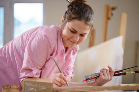 Cute woman paints on canvas in an art workshop. Artist creating picture. Art school or studio. Work with paints, brushes and easel. Hobby and leisure concept. Woman painter at workspace.の写真素材